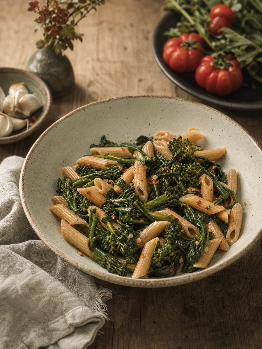 A wide, shallow ceramic bowl filled with whole wheat penne and dark green broccoli rabe, glistening with olive oil and speckled with red pepper flakes, set on a wooden table.
