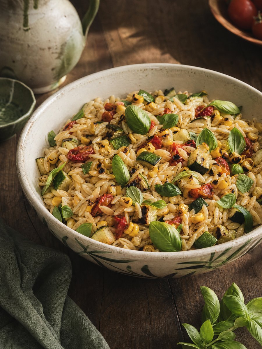 A large ceramic serving bowl filled with cooked orzo pasta, charred corn kernels, and diced grilled zucchini, garnished with fresh basil leaves on a wooden table.